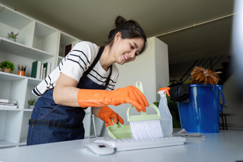 Asian woman with gloves cleaning keyboard with a small brush and pan, showcasing attention to detail and office maintenance.