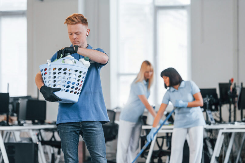 Woman uses vacuum cleaner. Group of workers clean modern office together at daytime.
